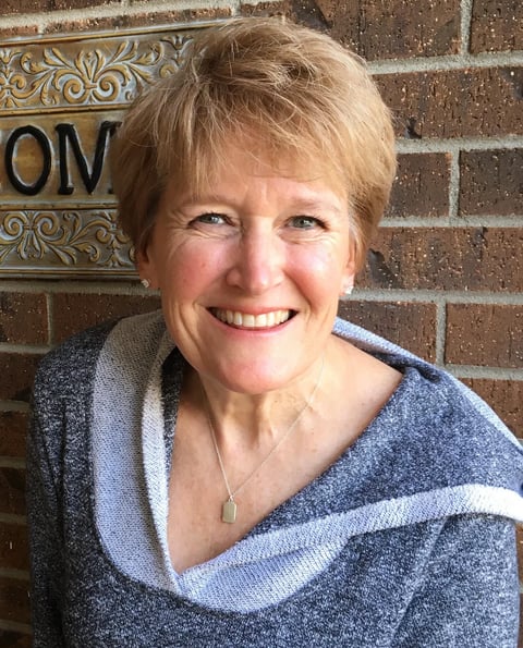 Smiling woman with short blonde hair wearing a gray and white striped sweater, photographed against a brick wall with a decorative gold sign