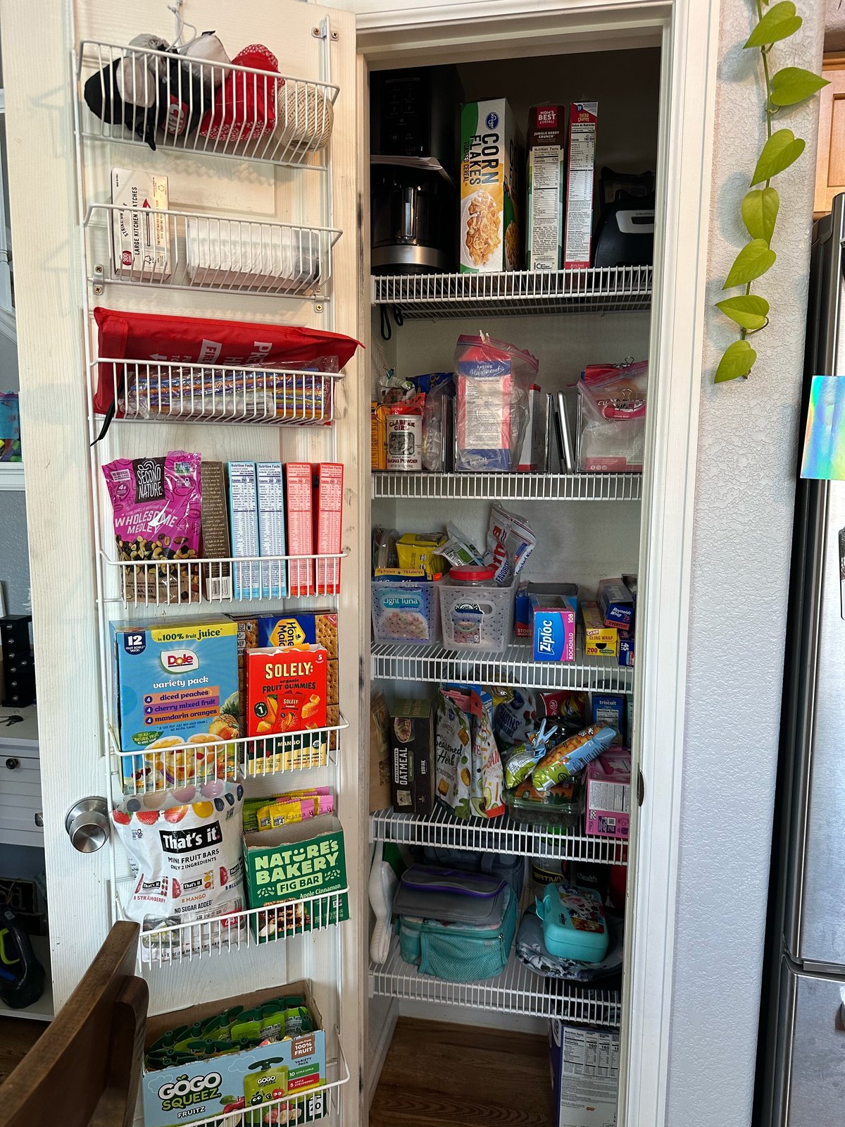 Organized kitchen pantry with white wire shelving and baskets storing food, snacks, and household items on a cabinet door