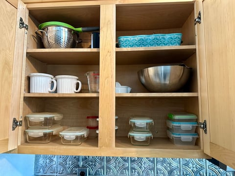 Open wooden kitchen cabinet displaying organized dishes, mugs, bowls, and food storage containers on multiple shelves